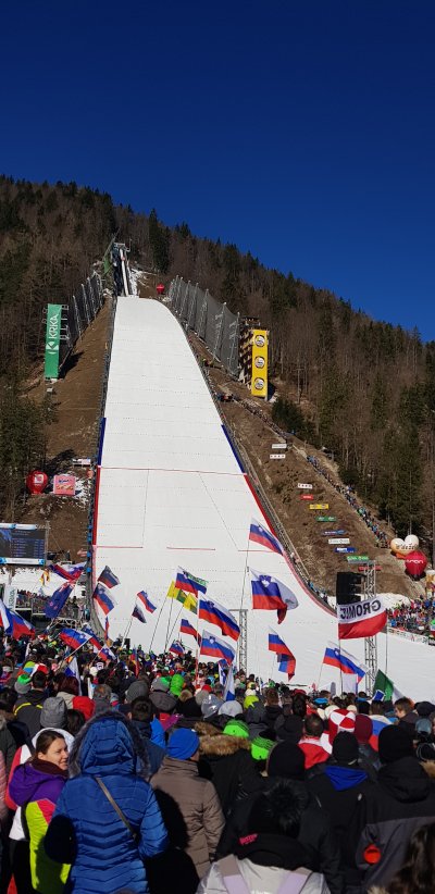 To je en tak pravi planiški dan, ki ga ne pozabiš.Mrzel zrak, sonce sije, okoli mene pa morje ljudi in slovenske zastave na vsakem koraku. V ozadju zasnežene gore, pred nami pa tisti poseben občutek, ko veš, da se bo nekaj velikega zgodilo. Vsi nasmejani, malo hripavi od navijanja, malo premraženi, ampak čisto naviti.Gužva, smeh, kakšna domača beseda z neznancem, ki v sekundi postane “naš”. Tisti občutek, ko si ponosen, ko te kar malo stisne pri srcu, ko množica zagrmi. Ni važno, od kod si – v Planici smo vsi isti.To ni samo tekma. To je dan za spomine. 